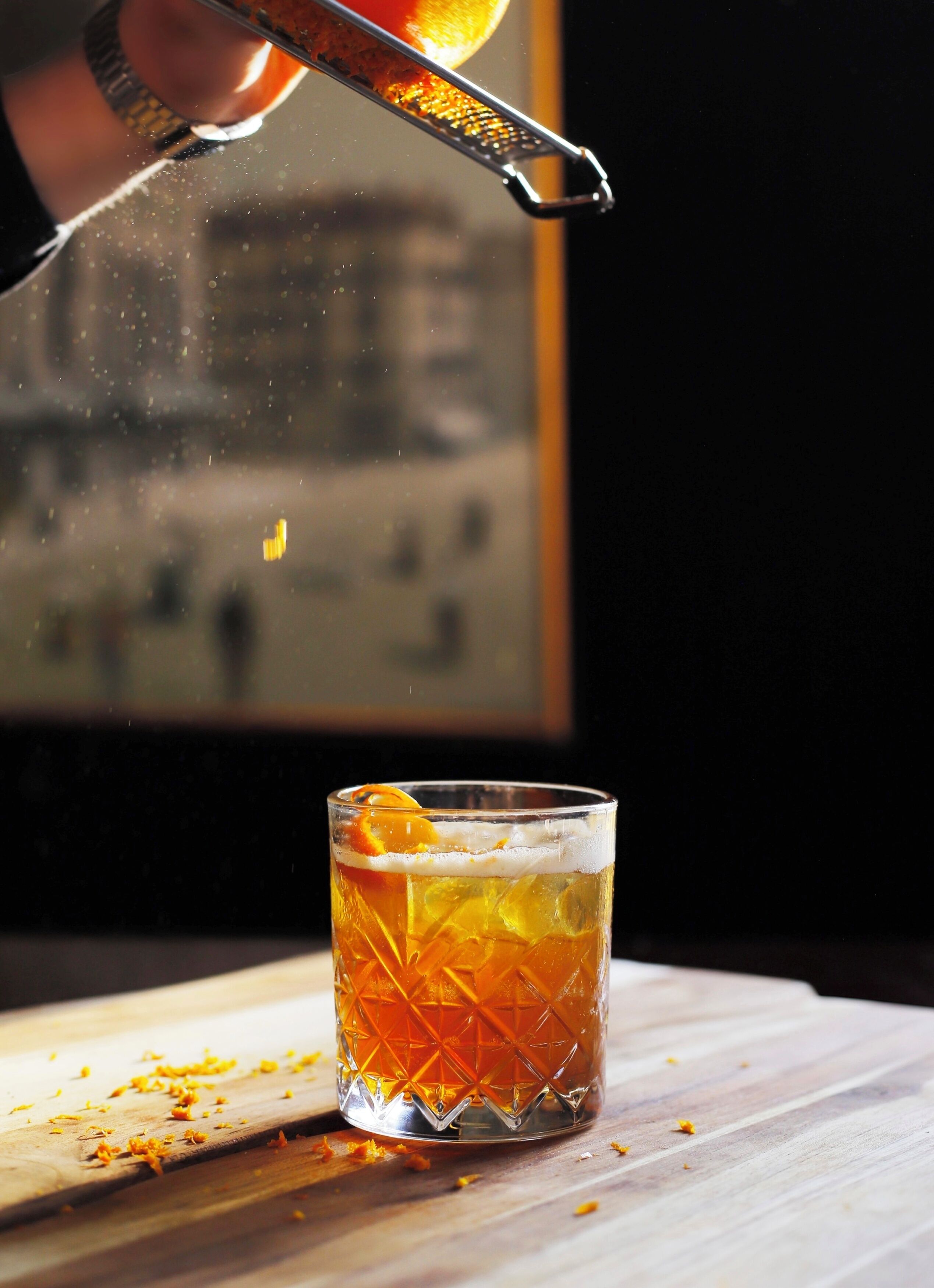 Un vaso de bebida color ámbar con hielo y una rodaja de naranja, sobre una superficie de madera con ralladura de naranja dispersa.