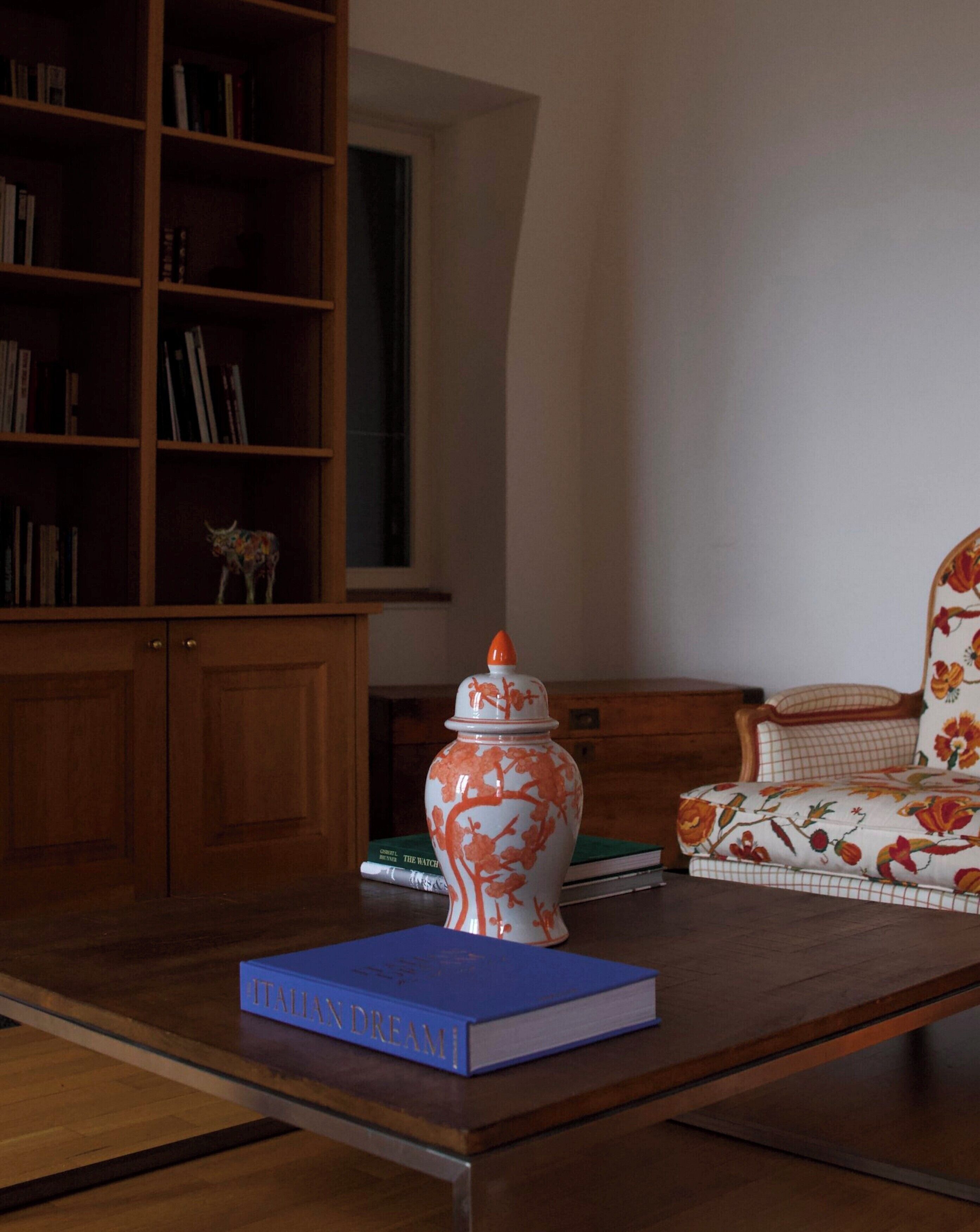 Ein Zimmer mit einem Holzbücherregal, einer dekorativen Keramikvase, einem blauen Buch mit dem Titel 'ITALIAN DREAM' und einem armchair mit Blumenmuster.