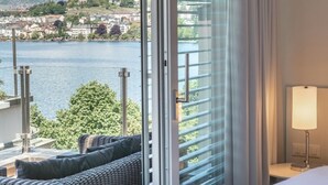 A hotel room with a large sliding glass door leading to a balcony with a view of a lake and distant buildings. - Rouvenaz Appartements (Montreux)
