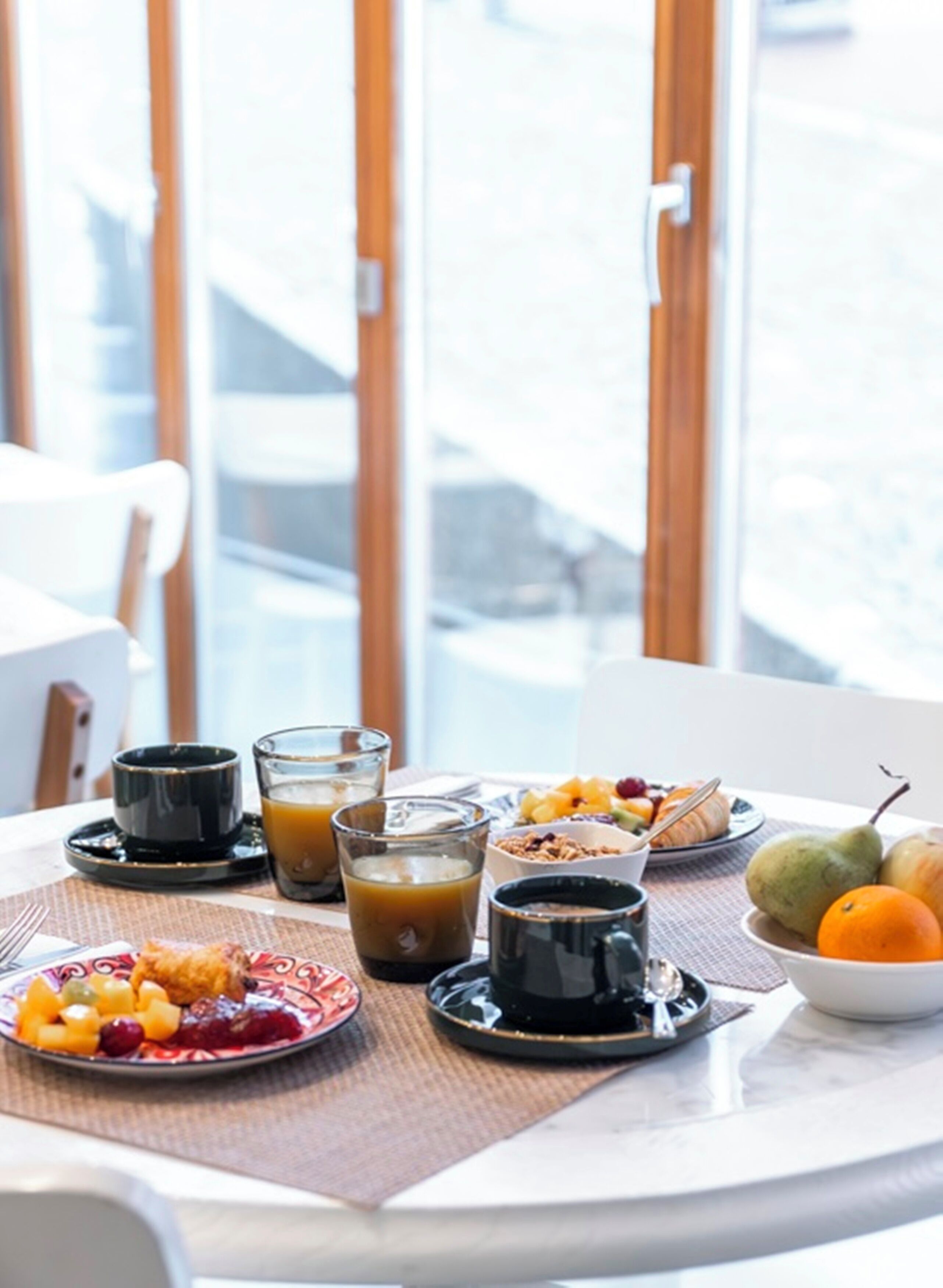Una mesa de desayuno con una variedad de alimentos, incluyendo frutas, un croissant y bebidas en tazas negras.