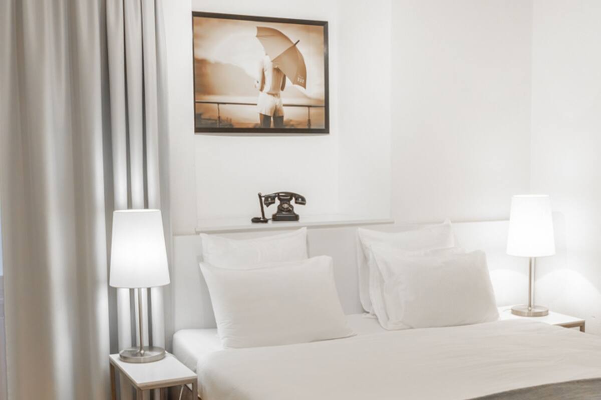 Una habitación de hotel con cama, dos mesitas de noche con lámparas, un cuadro en la pared y una ventana con cortinas.