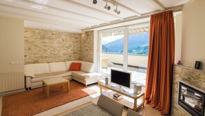 A modern living room with a stone wall, a white sofa, a flat-screen TV, a fireplace, and a large window with a mountain view. - The Cristal apartment house is situated in a quiet, sunny and central location (Leukerbad)