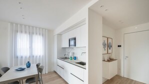 A modern kitchen with white cabinets, a dining table with chairs, and a window with sheer curtains. - Prestige and Comfort in Lignano (Lignano Sabbiadoro)