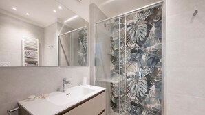 Un baño moderno con un tocador blanco, un espejo grande y una ducha con puerta de vidrio que presenta un diseño de hojas tropicales.