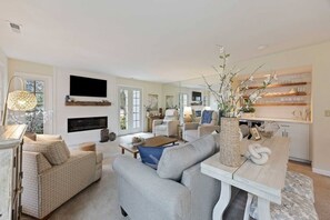 A modern living room with a fireplace, comfortable seating, and a kitchen area visible through an open door.