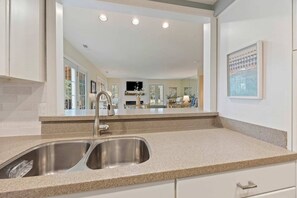 A kitchen with a double sink, granite countertop, and a view into the living area through a large mirror.