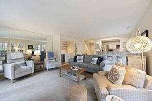 Een ruime woonkamer met een bank, fauteuils en een salontafel. In de achtergrond is een keuken te zien.