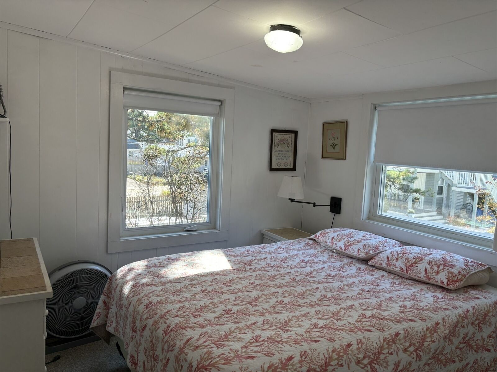 Un dormitorio con cama, dos ventanas, una mesita de noche, una lámpara y un cuadro en la pared.