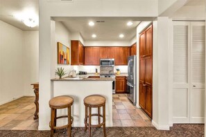 A kitchen with wooden cabinets, a central island with stools, a microwave, and a refrigerator.