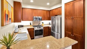 A modern kitchen with wooden cabinets, stainless steel appliances, granite countertops, and a potted plant. - Desert Bloom A | 1BR Legacy Villa (La Quinta)