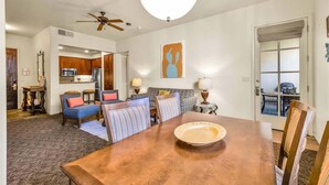 A spacious living room with a wooden dining table, a ceiling fan, and a kitchen visible through an open door. - Desert Bloom A | 1BR Legacy Villa (La Quinta)