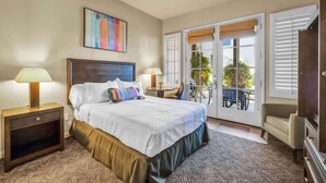 A bedroom with a bed, two bedside lamps, a ceiling fan, a painting, and a balcony with a table and chairs. - Desert Bloom A | 1BR Legacy Villa (La Quinta)