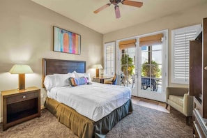Une chambre à coucher comprenant un lit, deux lampes de chevet, un ventilateur de plafond, un tableau et un balcon avec une table et des chaises.