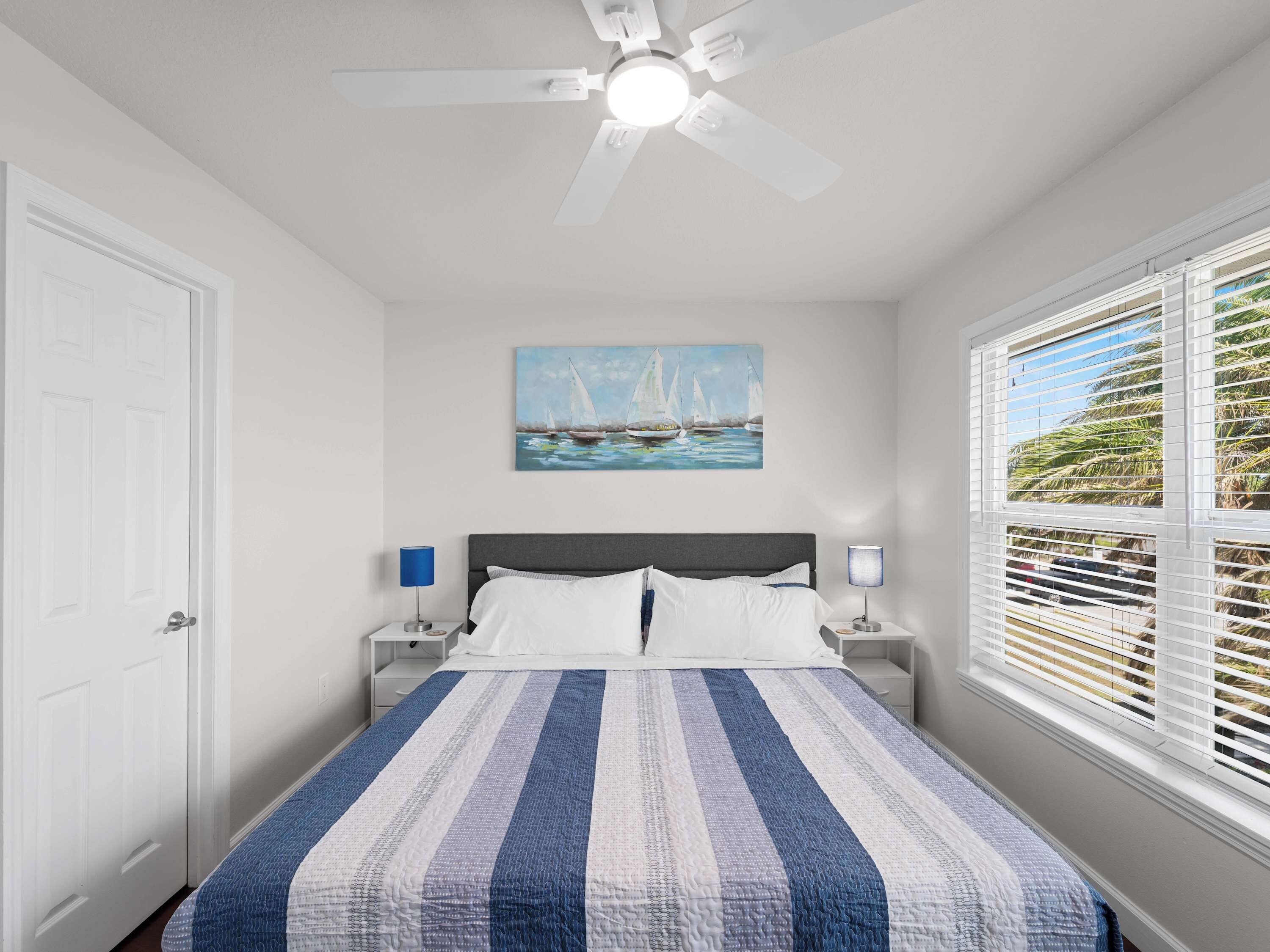 Une chambre à coucher comprenant un lit, deux tables de chevet, un ventilateur de plafond, un tableau représentant des voiliers et une fenêtre avec des stores.