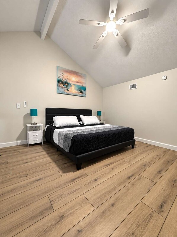 A bedroom with a bed, nightstands, a ceiling fan, a painting, and a smoke detector.