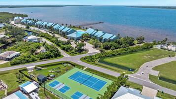 Une vue aérienne d’un hôtel situé sur la côte, avec plusieurs bâtiments aux toits bleus, un court de tennis et une jetée qui s’avance dans l’eau.