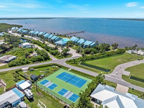 An aerial view of a coastal hotel with multiple blue-roofed buildings, a tennis court, and a pier extending into the water.