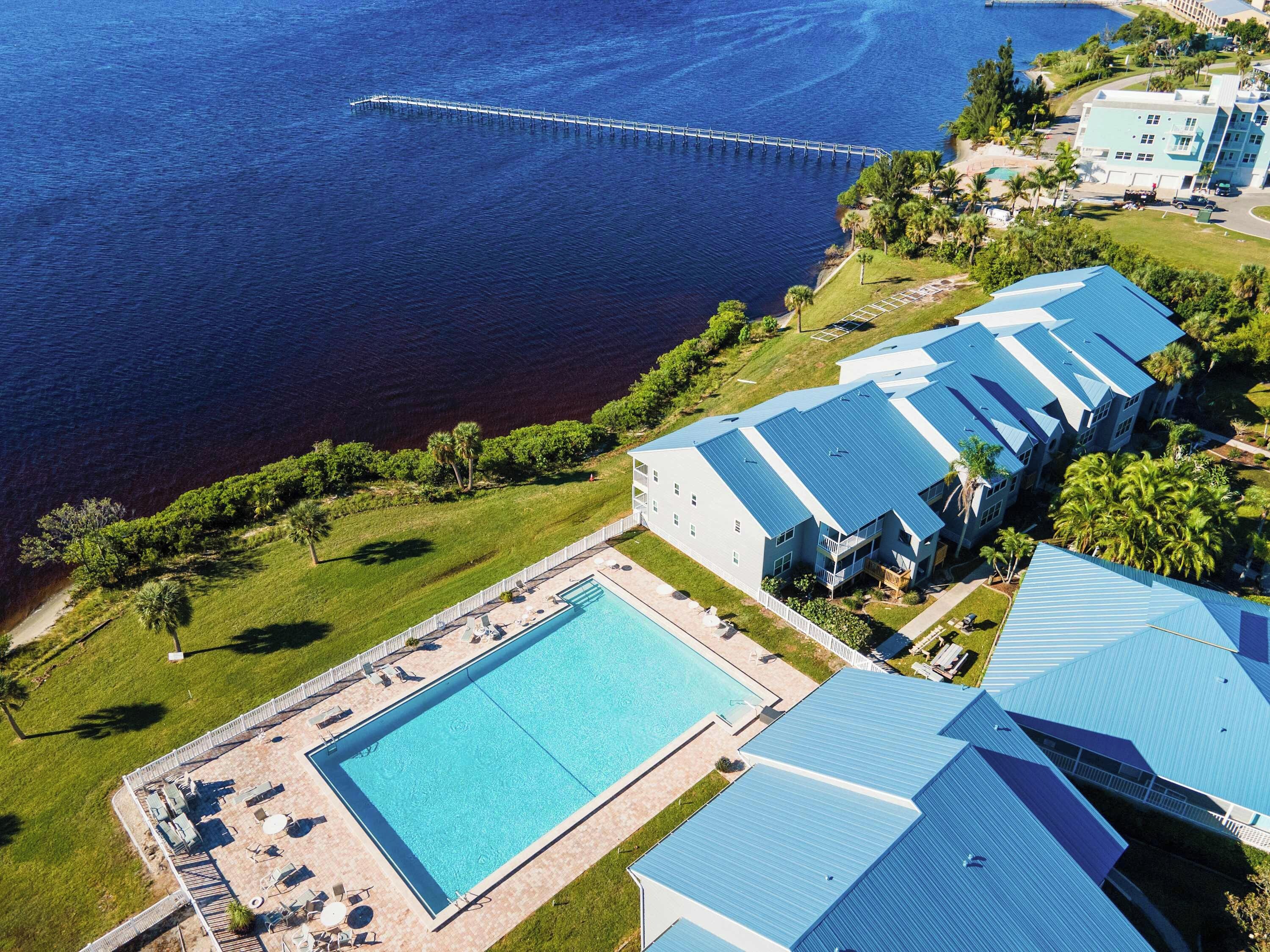 Une vue aérienne d’un complexe hôtelier avec une grande piscine, plusieurs bâtiments aux toits bleus et une jetée qui s’avance dans l’eau.