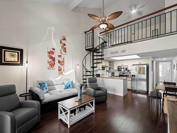 A modern living room with a grey sofa, a coffee table, and a dining area with a wooden table and chairs. A kitchen is visible in the background with a stainless steel refrigerator and microwave.