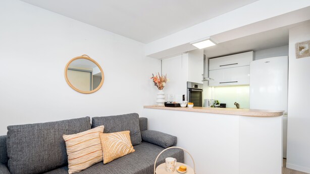 Un espace de vie compact comprenant un canapé gris, une petite table ronde avec une tasse et une assiette, et une kitchenette dotée de meubles blancs et d’un réfrigérateur.