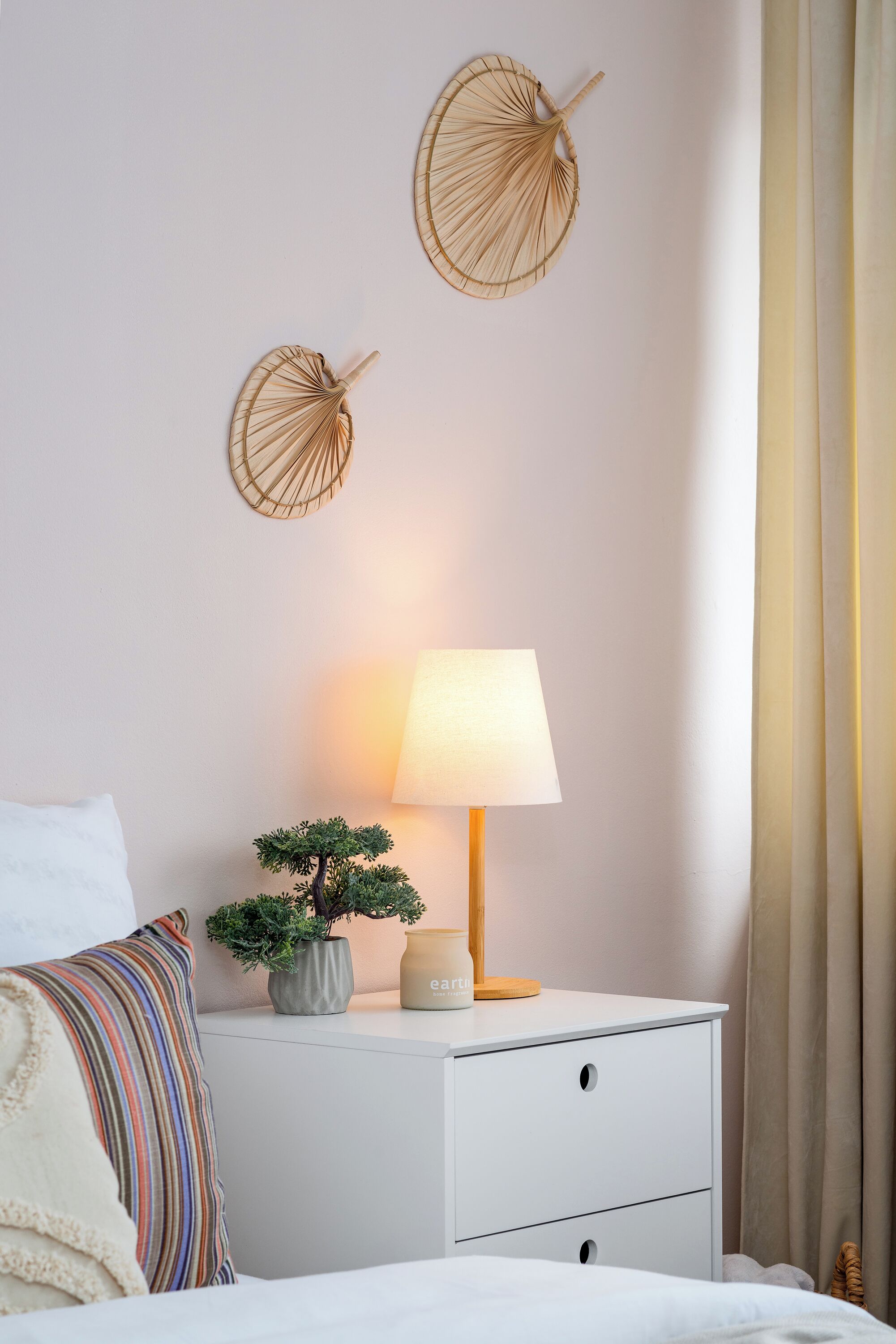 Una camera da letto ordinata con un comodino bianco, una lampada da comodino, un piccolo vaso con una pianta e due ventole decorative a parete.