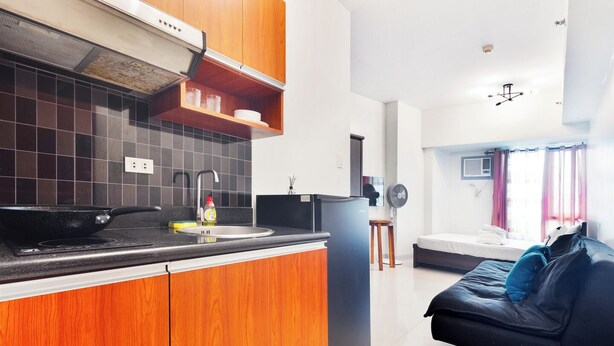 A modern kitchen with wooden cabinets, a black countertop, and a built-in oven. There is a dining area with a black sofa and a small table. - Cozy studio condo with WiFi, AC in beautiful Makati (Makati)