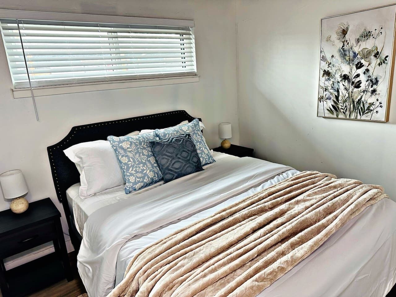 A bedroom with a bed, a nightstand, a lamp, a window with blinds, and a wall-mounted picture.