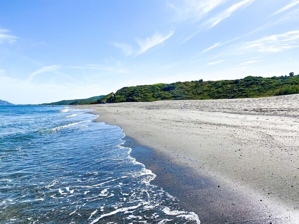 A beach with clear blue water, a sandy shore, and a green hillside in the background.