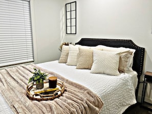 Una cama bien hecha con cabecera, una mesita de noche, una ventana con persianas y una planta junto a la cama.