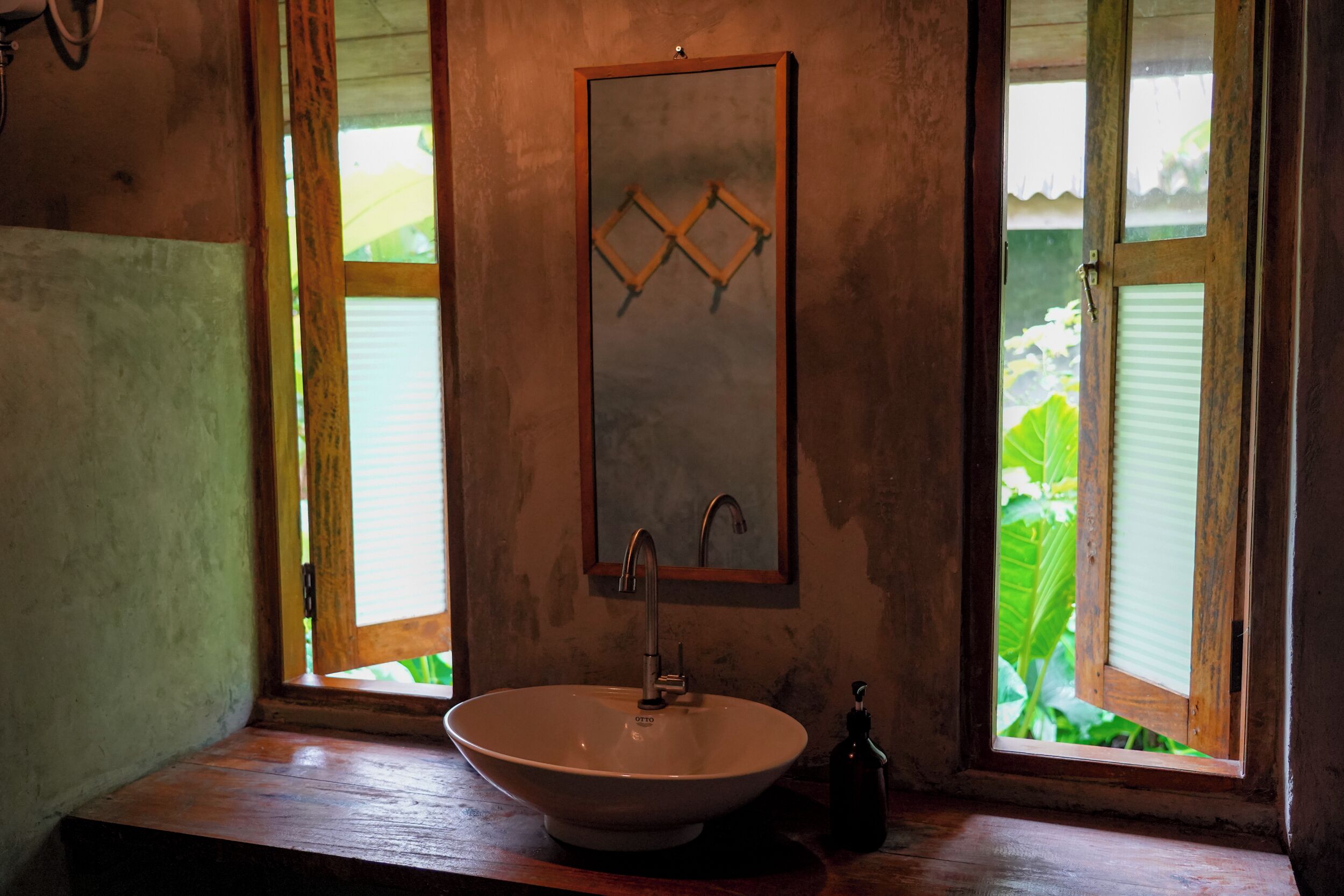 Une salle de bain avec un meuble-lavabo en bois, un lavabo sur colonne, un miroir et une fenêtre donnant sur un paysage verdoyant.