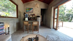 A cozy room with a wooden ceiling, a dining area with a round table and chairs, a kitchen area with a fridge and a water dispenser, and a large window with a view of greenery. - Into The Blue Sri Lanka (Tangalle)