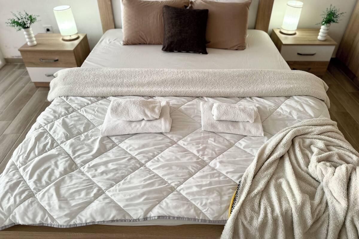 A neatly made bed with a quilted duvet, pillows, and a blanket. There are bedside tables with lamps and small potted plants.