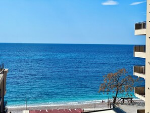 Vista costera con cielo despejado, una playa y un edificio con balcones que dan al mar.