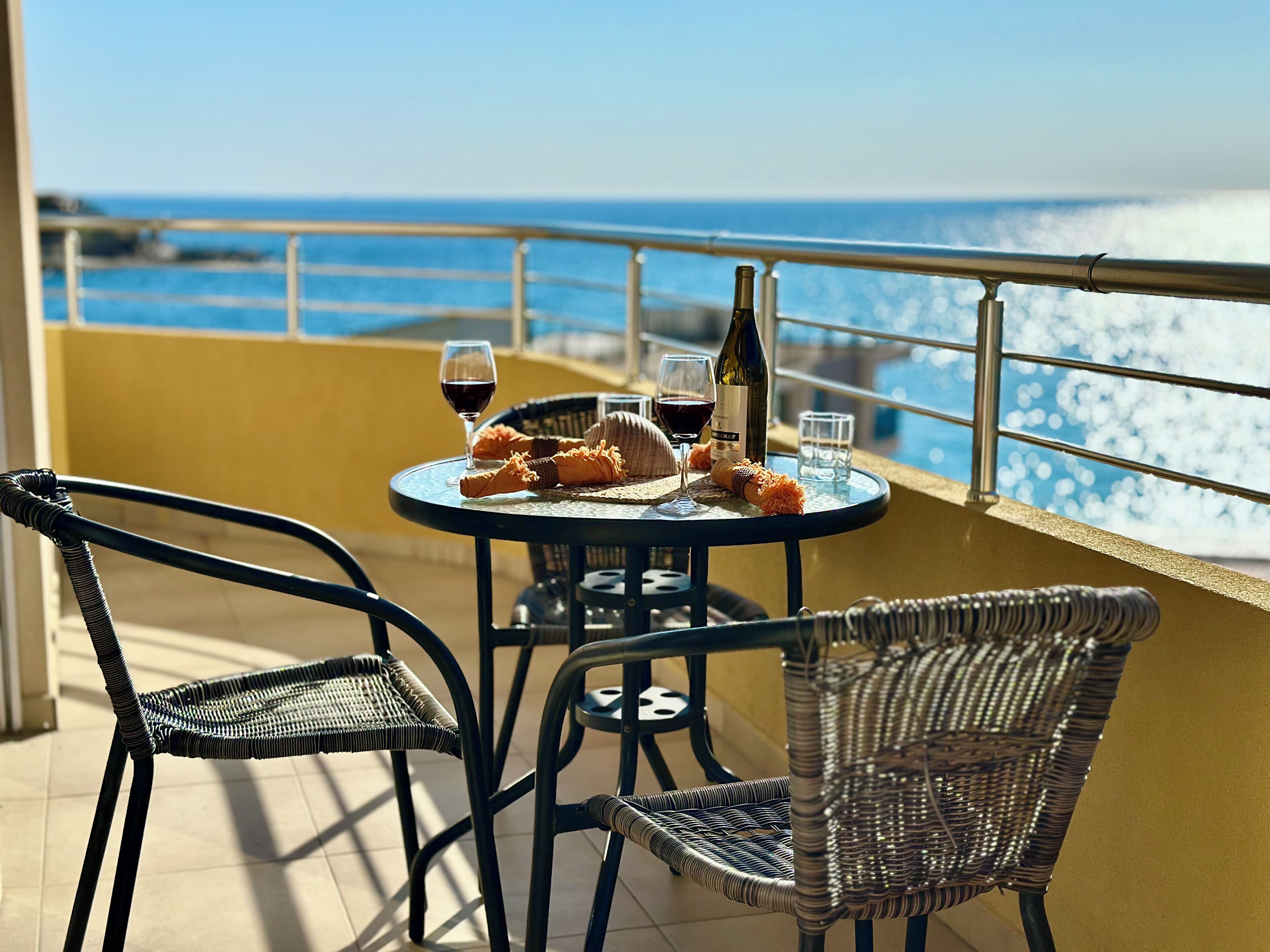 A round table with a bottle of wine, glasses, and bread on a balcony overlooking the sea.