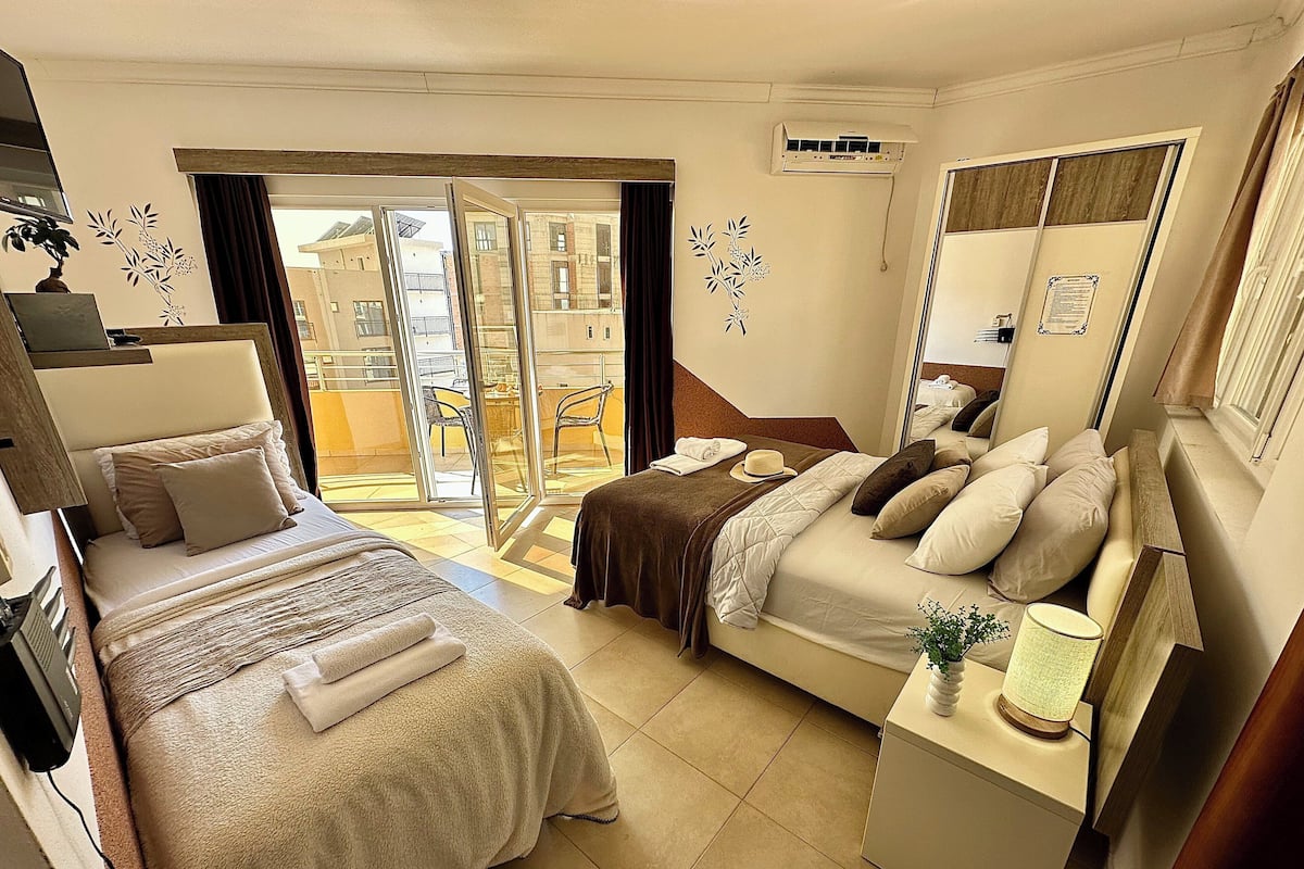 Habitación de hotel con dos camas, un escritorio, una silla, una mesita de noche con lámpara y una puerta corrediza de vidrio que da a un espacio exterior.