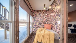 Une chambre avec une grande fenêtre, un lit recouvert d’une couverture jaune, un papier peint à motifs floraux et un plancher en bois.