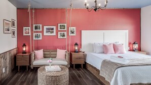 Un dormitorio con una pared roja, un cabecero blanco, dos mesitas de noche con faroles, una cama con ropa de cama blanca y un reposapiés de mimbre.