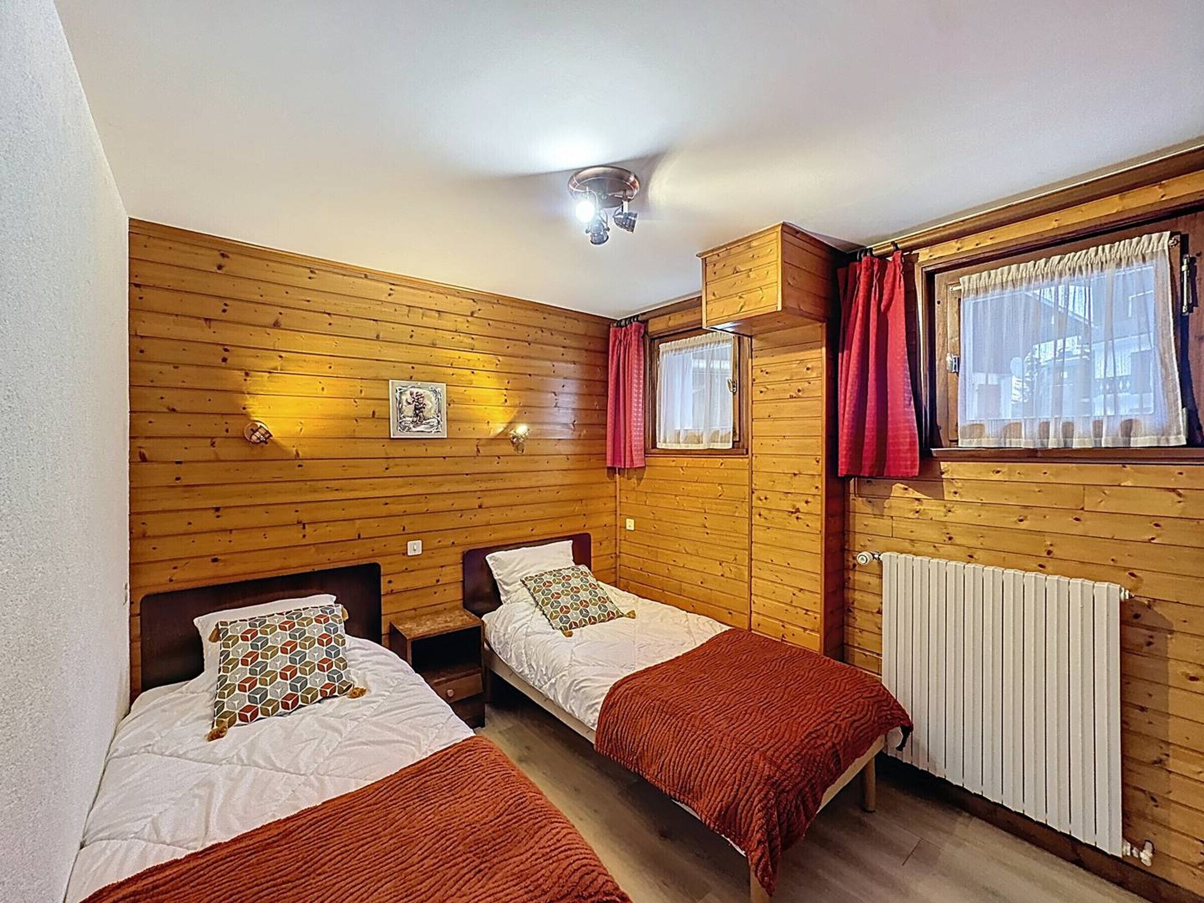 Une chambre avec deux lits, des murs en bois, un luminaire au plafond, une fenêtre avec des rideaux et un radiateur.