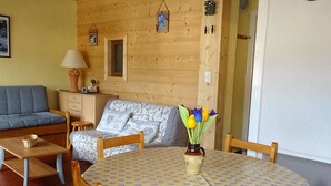 Une pièce chaleureuse avec un mur en bois, un canapé, une table à manger recouverte d’une nappe à motifs et un vase rempli de fleurs.