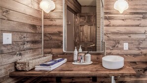 A wooden cabin bathroom with a large mirror, two wall sconces, a sink, and toiletries.