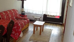 Un saló amb un sofà, una taula petita, una cadira, un televisor fixat a la paret i una finestra amb cortines.
