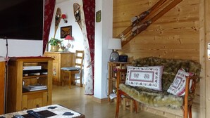 A cozy cabin interior with wooden walls, a sofa with fur throw, a dining area with a table and chairs, and skis leaning against the wall.