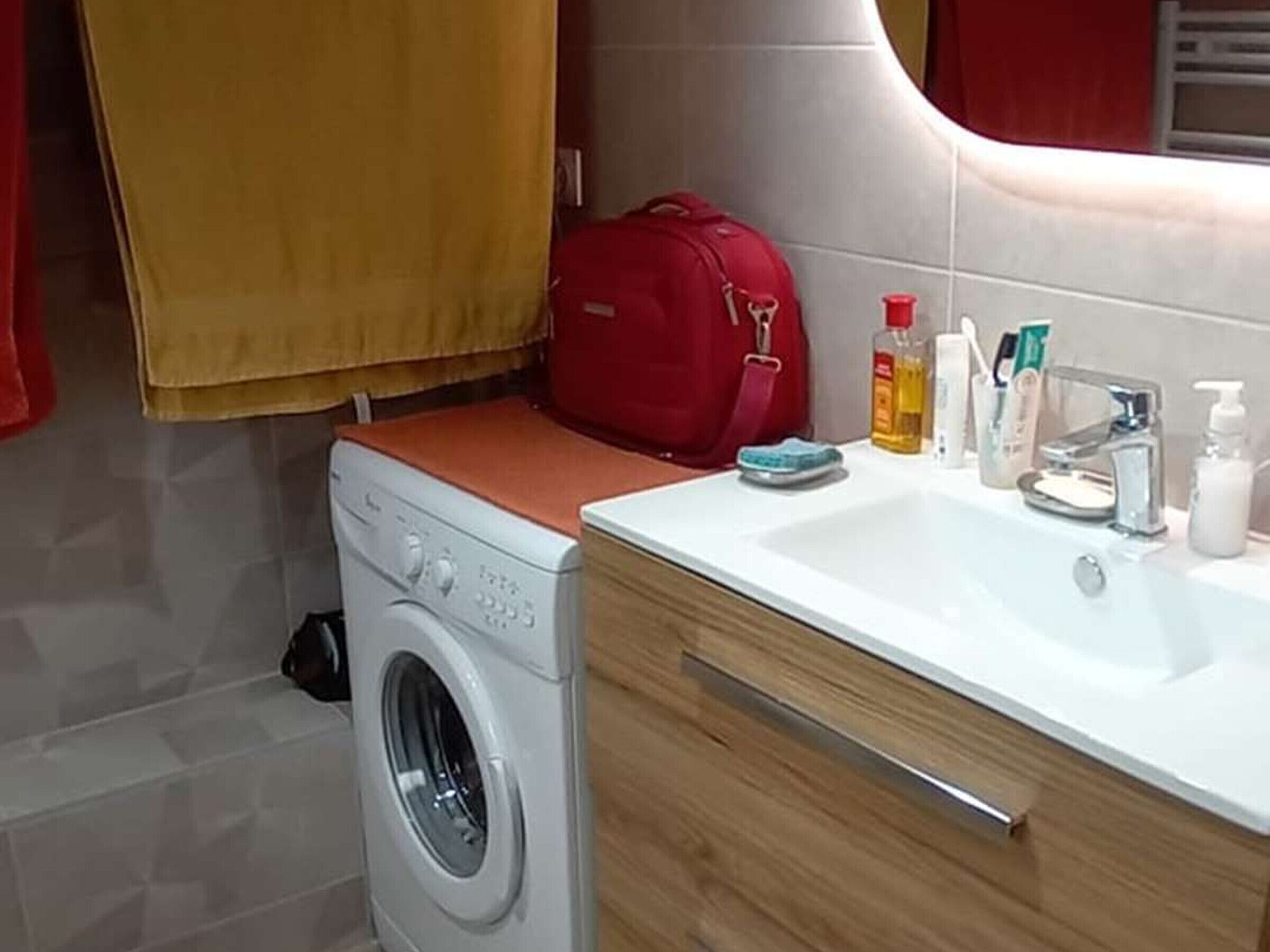 A compact bathroom with a washing machine, a sink with toiletries, and a red bag on the counter.