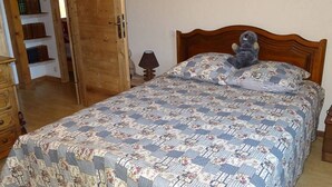 A bedroom with a wooden bed, floral-patterned bedding, a teddy bear on the bed, and a wooden wardrobe with shelves.