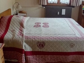 Ein ordentlich bezogenes Bett mit einer gesteppten Decke mit Herzmuster, einer Nachttischlampe und einem Fenster mit Vorhängen.