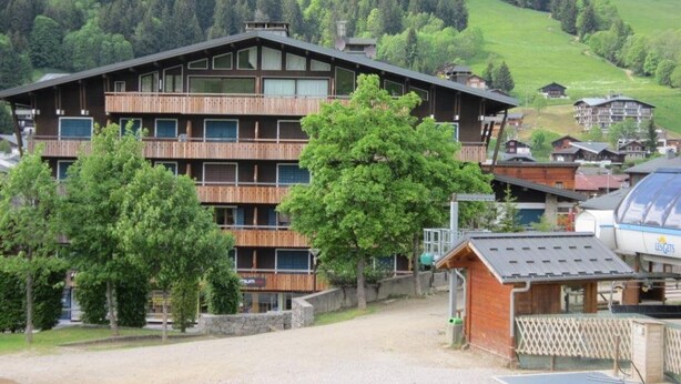 A multi-story wooden building with balconies, surrounded by trees and a hilly landscape.