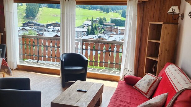 A living room with a red sofa, a black chair, and a wooden coffee table. A balcony with a wooden railing and a view of a village is visible through the window. - 4-bedroom apartment for 8 with garage, near shops and cable cars (Les Gets)