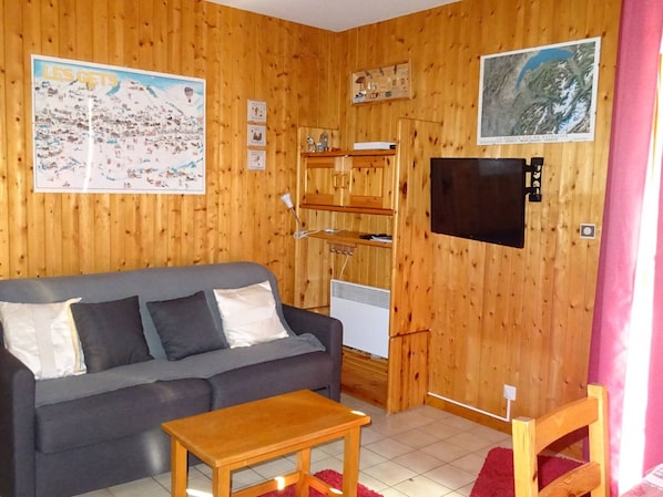 A cozy room with a wooden wall, a grey sofa, a wooden table, and a TV mounted on the wall.