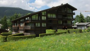 A multi-story wooden building with balconies, surrounded by well-maintained hedges and a mountainous backdrop.
