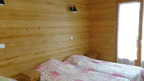 En stuga med träpanel, inredd med två sängar, ett litet träbord som sängbord, en vägglampa och ett fönster med gardin.